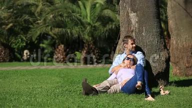 一对浪漫的情侣坐在棕榈树下。 一个女孩在一个男人的大腿上。 一对相爱的夫妇在草地上的公园里休息